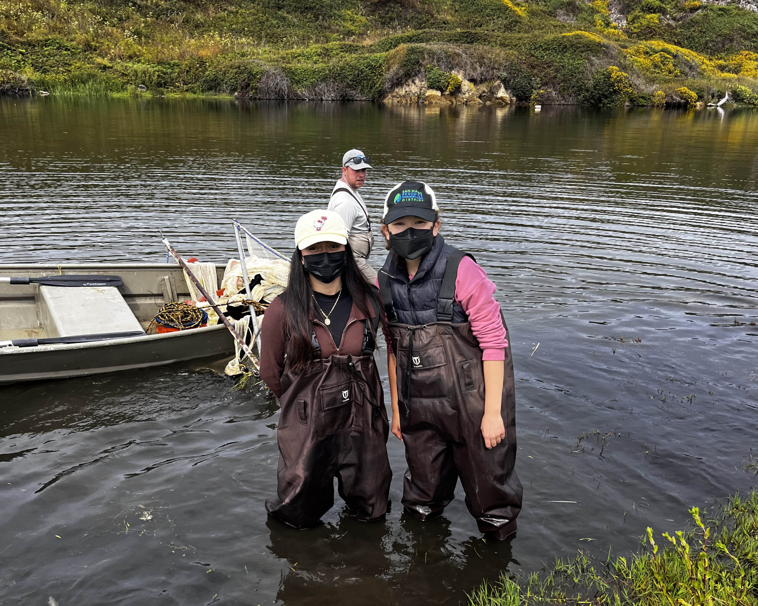 Nature's stewards Two people standing together for a photo in a lake in waders and masks, with a rowboat and a third person behind them.