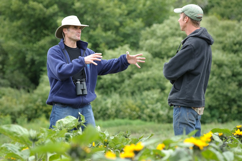 NRCS partnership Outside, a person with binoculars and a brimmed had gesturing with both hands as he speaks to another person.